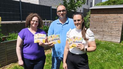 Drei Personen präsentieren im Garten die gelb‑weißen Flyer „HELLWEG SOMMER 2026“ vor einem blühenden Beet und einem Holzschuppen.